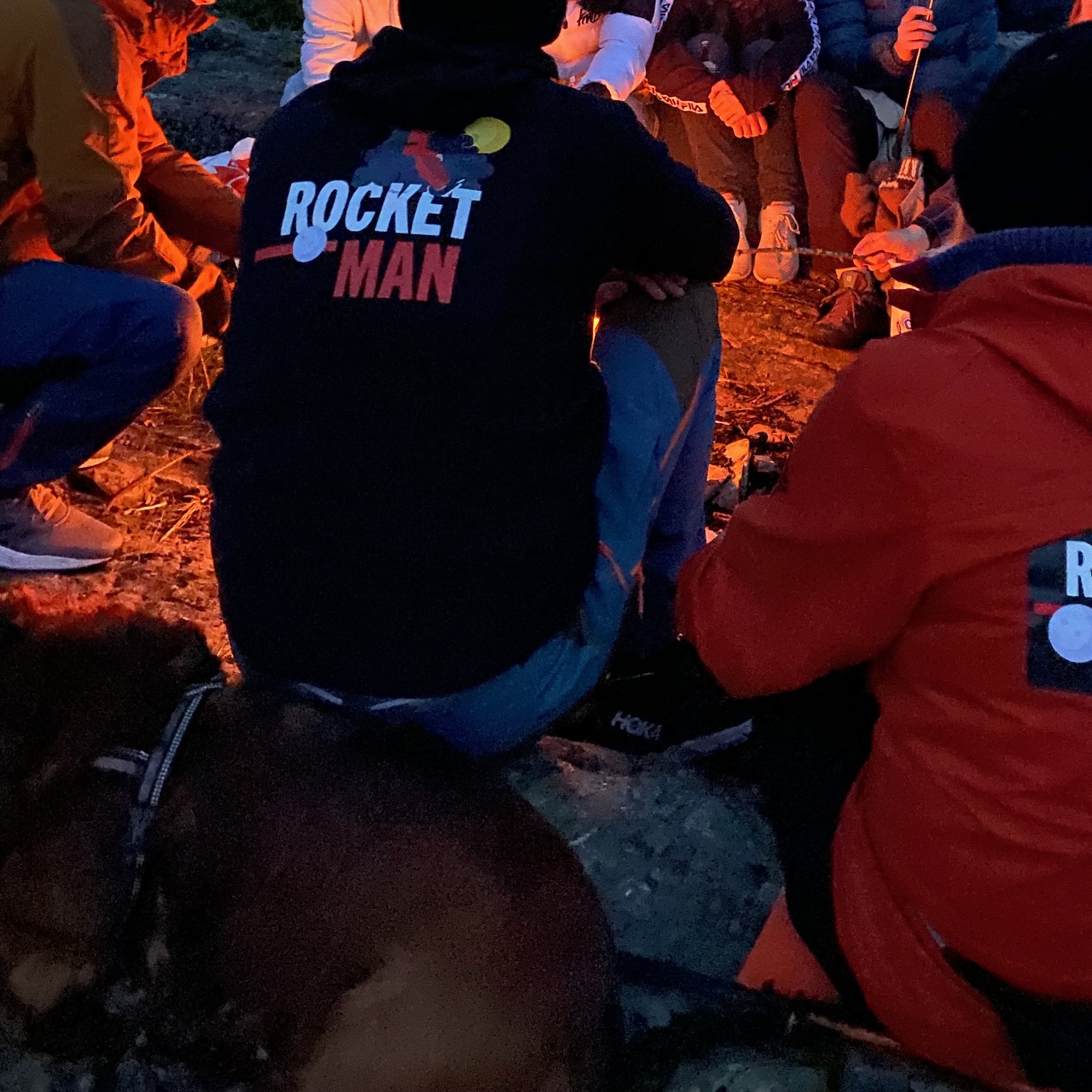 Mennsker som sitter rundt et bål. En har en genser med påskriften "Rocket Man"